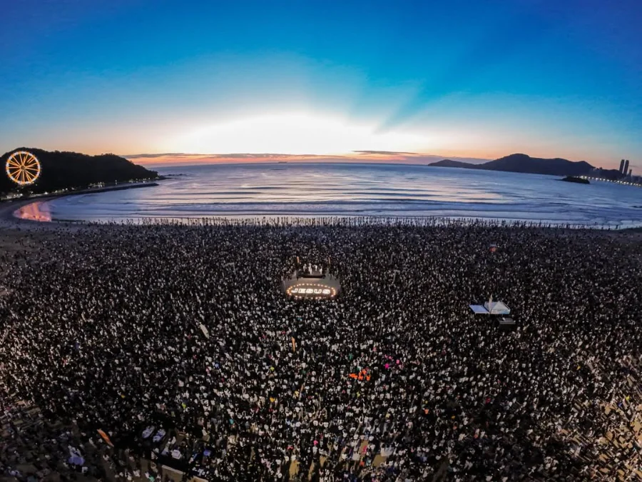 BC 15 Mil Pessoas Oram ao Amanhecer na Sexta-Feira Santa Vista aérea de multidão reunida na areia da Praia Central de Balneário Camboriú durante o evento Epiphany Movement ao amanhecer da Sexta-feira Santa. Ao centro, um letreiro iluminado escrito JESUS e a roda-gigante FG Big Wheel ao fundo à esquerda sob o céu do nascer do sol.