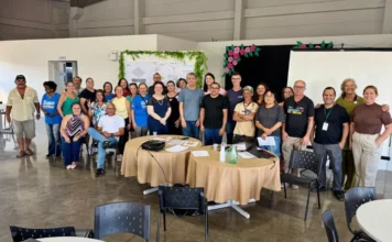 Balneário Arroio do Silva sedia 2ª etapa da Jornada CRIATUR Participantes e organizadores posando para foto de grupo durante a segunda etapa da Jornada CRIATUR de Inovação em Turismo e Economia Criativa, no Centro Multiuso Esther Pacheco Mucillo em Balneário Arroio do Silva. O fundo mostra um quadro de co-criação com hera verde e decorações de flores.