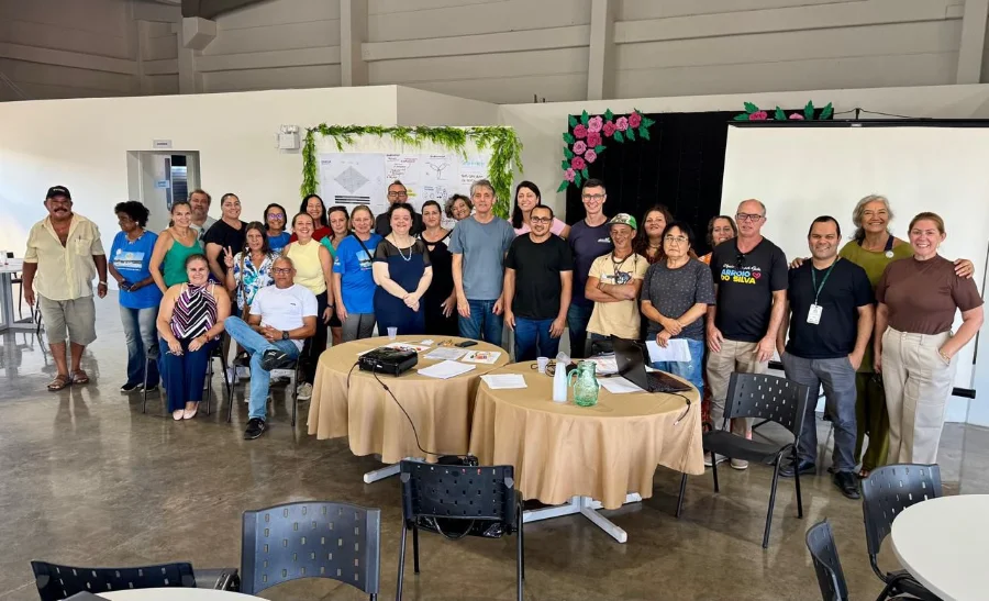 Participantes e organizadores posando para foto de grupo durante a segunda etapa da Jornada CRIATUR de Inovação em Turismo e Economia Criativa, no Centro Multiuso Esther Pacheco Mucillo em Balneário Arroio do Silva. O fundo mostra um quadro de co-criação com hera verde e decorações de flores.