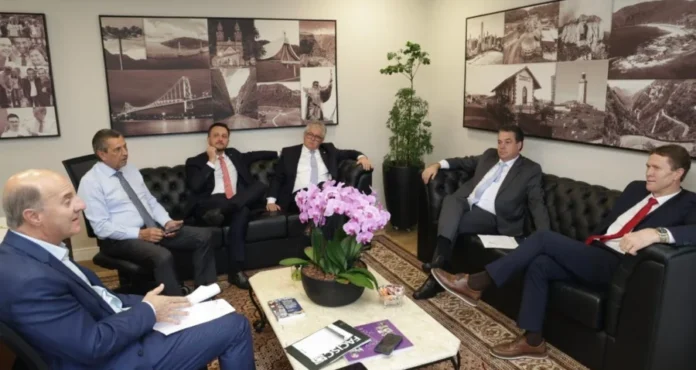 Bancada do Sul debate Rio Urussanga e custo do gás na Alesc Deputados estaduais da Bancada do Sul reunidos em gabinete na Alesc para debater o desassoreamento do Rio Urussanga e o custo do gás natural. Na cena, parlamentares conversam sentados em sofás pretos ao redor de uma mesa com orquídeas e documentos.