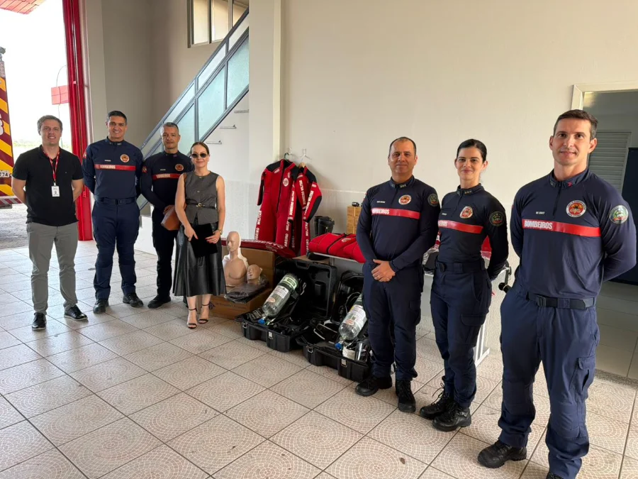 Entrega simbólica de novos equipamentos operacionais e de prevenção para o Corpo de Bombeiros de Forquilhinha (CBMSC) em parceria com o MPSC. A promotora de Justiça Rafaela Póvoas Cardozo Lehmann e oficiais posam com os materiais doados, incluindo manequins de RCP, Equipamentos de Proteção Respiratória (EPRs), capacetes e roupas de neoprene, no interior de um quartel.
