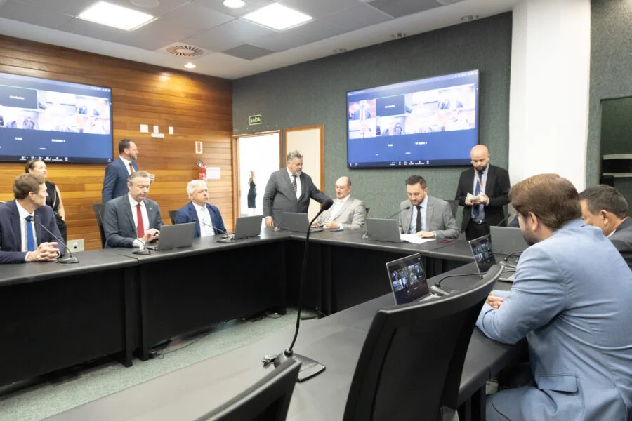 Deputados reunidos em sessão extraordinária da CCJ em mesa redonda com notebooks e telas de videoconferência para votação da MP 269/2026 sobre a descompactação da tabela salarial dos professores estaduais.