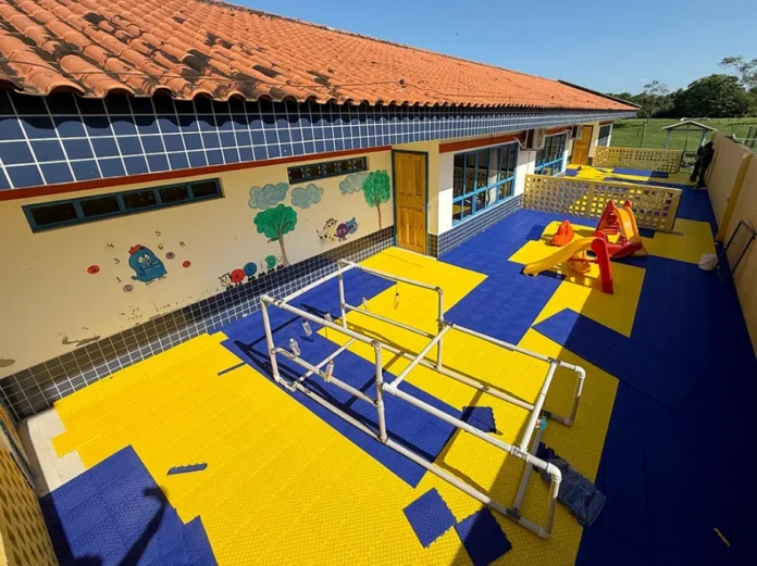 CEI de Santa Rosa do Sul instala novos pisos para crianças Área externa do CEI Vovó Leoncina em Santa Rosa do Sul exibindo a instalação de piso modular colorido nas cores azul e amarelo, com brinquedos de parquinho e estrutura pedagógica de PVC para atividades infantis.