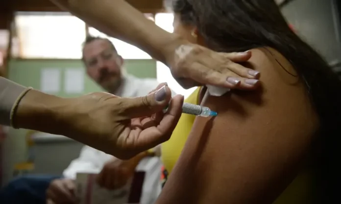 CLT obriga empresas a informar sobre câncer e vacinas Close-up focado no braço de uma mulher recebendo uma vacinação no deltoide. As mãos de profissionais de saúde aplicam a injeção e um algodão. Um médico desfocado observa ao fundo em um consultório, ilustrando o processo de imunização e prevenção em saúde.