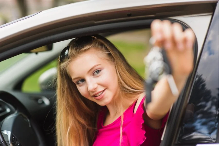 CNH para menores Câmara discute redução da idade mínima Jovem loira sorrindo no banco do motorista de um carro, estendendo as chaves pela janela. Cena ilustrativa sobre o debate de redução da idade mínima para CNH.