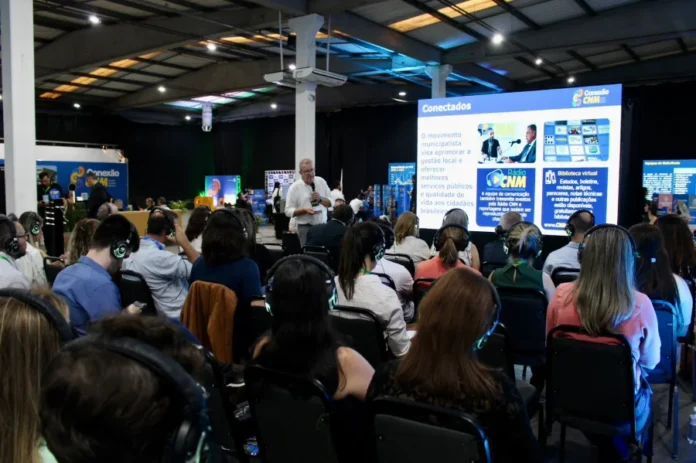 CNM reúne mil gestores em Criciúma para qualificar gestão Público de gestores e servidores municipais utilizando fones de ouvido durante palestra na imersão Conexão CNM em Criciúma. Ao fundo, palestrante apresenta painel sobre ferramentas de gestão pública municipal e recursos da Confederação Nacional de Municípios.