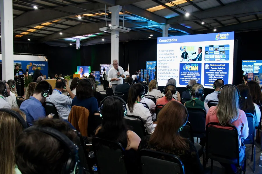 Público de gestores e servidores municipais utilizando fones de ouvido durante palestra na imersão Conexão CNM em Criciúma. Ao fundo, palestrante apresenta painel sobre ferramentas de gestão pública municipal e recursos da Confederação Nacional de Municípios.