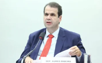 Câmara aprova avisos de riscos à saúde em celulares O deputado federal Aureo Ribeiro, relator do projeto, fala ao microfone durante sessão na Câmara dos Deputados. Ele veste paletó azul e gravata vermelha, segurando documentos sobre a mesa. Ao fundo, uma parede clara e a placa de identificação com seu nome. Foco em acessibilidade e SEO para termos como legislação e direitos do consumidor.