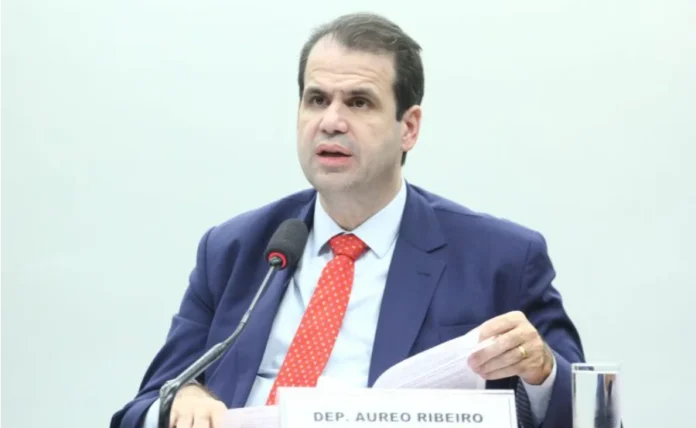 Câmara aprova avisos de riscos à saúde em celulares O deputado federal Aureo Ribeiro, relator do projeto, fala ao microfone durante sessão na Câmara dos Deputados. Ele veste paletó azul e gravata vermelha, segurando documentos sobre a mesa. Ao fundo, uma parede clara e a placa de identificação com seu nome. Foco em acessibilidade e SEO para termos como legislação e direitos do consumidor.