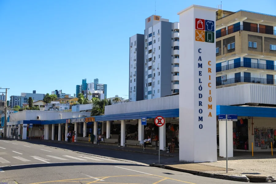 Fachada externa do Camelódromo de Criciúma em um dia ensolarado, destacando o totem vertical de identificação e a estrutura azul do centro de compras. A imagem ilustra a abertura do novo edital para ocupação de espaços comerciais no local.