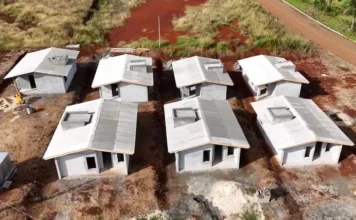 Casa Catarina: Primeiras casas serão entregues em breve Vista aérea de canteiro de obras com sete moradias populares em construção do programa habitacional Casa Catarina, em Santa Catarina. As casas estão em estágio avançado, com paredes e telhado prontos, em meio a solo vermelho e materiais de construção. Foto ilustrativa do progresso das obras.