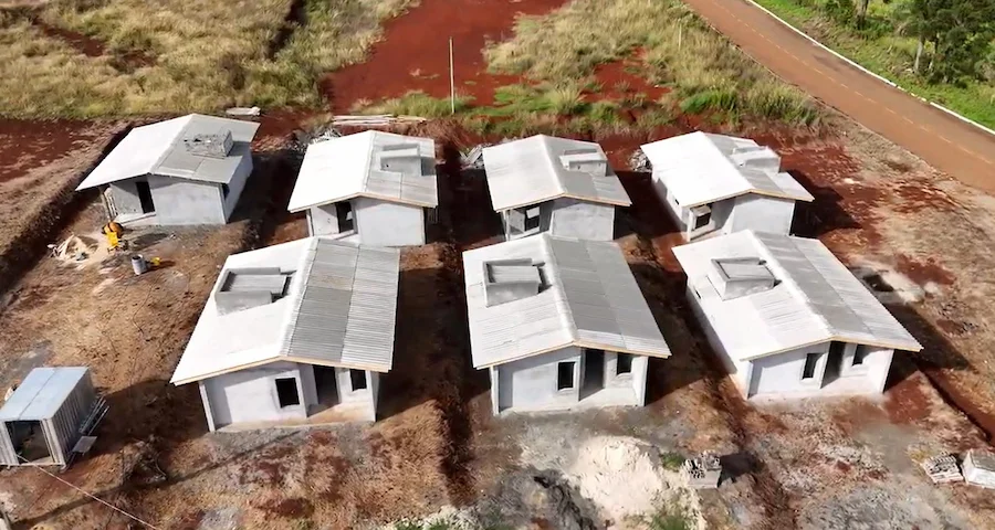 Vista aérea de canteiro de obras com sete moradias populares em construção do programa habitacional Casa Catarina, em Santa Catarina. As casas estão em estágio avançado, com paredes e telhado prontos, em meio a solo vermelho e materiais de construção. Foto ilustrativa do progresso das obras.