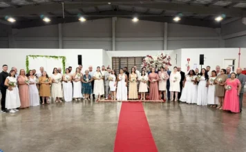 Casamento coletivo une 21 casais em Arroio do Silva Vinte e um casais reunidos em fileira durante a cerimônia de Casamento Coletivo 2026 em Balneário Arroio do Silva. A imagem mostra os noivos e noivas em trajes formais em um salão com tapete vermelho e decoração floral ao fundo.
