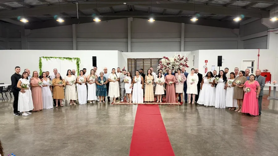 Vinte e um casais reunidos em fileira durante a cerimônia de Casamento Coletivo 2026 em Balneário Arroio do Silva. A imagem mostra os noivos e noivas em trajes formais em um salão com tapete vermelho e decoração floral ao fundo.