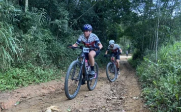Cicloturismo de Morro da Fumaça reúne mais de 300 atletas Dois ciclistas de montanha (MTB) com uniformes da equipe MATTOS pedalando em uma trilha de estrada de terra cercada por floresta densa em Morro da Fumaça, Santa Catarina, durante o percurso Sport desafiador da 6ª edição do Cicloturismo. A ciclista da frente veste capacete azul e tem o número de peito 849. O segundo ciclista segue atrás em uma bicicleta preta e vermelha.