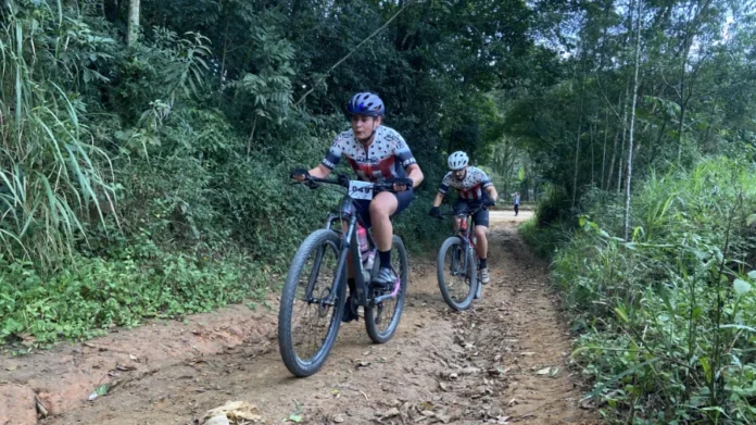 Cicloturismo de Morro da Fumaça reúne mais de 300 atletas Dois ciclistas de montanha (MTB) com uniformes da equipe MATTOS pedalando em uma trilha de estrada de terra cercada por floresta densa em Morro da Fumaça, Santa Catarina, durante o percurso Sport desafiador da 6ª edição do Cicloturismo. A ciclista da frente veste capacete azul e tem o número de peito 849. O segundo ciclista segue atrás em uma bicicleta preta e vermelha.