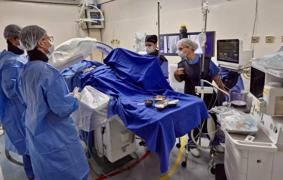 Profissionais de saúde realizam procedimento de hemodinâmica no Instituto de Cardiologia de Santa Catarina (ICSC). A imagem detalha a equipe médica com trajes cirúrgicos operando equipamentos de alta tecnologia e monitores em uma sala de cirurgia cardiovascular moderna.
