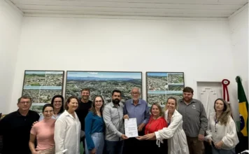 Cocal do Sul investe R$ 4,3 milhões em obras e habitação Prefeito Ademir Magagnin e autoridades de Cocal do Sul seguram documento de ordem de serviço para pacote de obras de R$ 4,3 milhões em habitação e infraestrutura. Ao fundo, mapas aéreos da cidade em gabinete municipal.