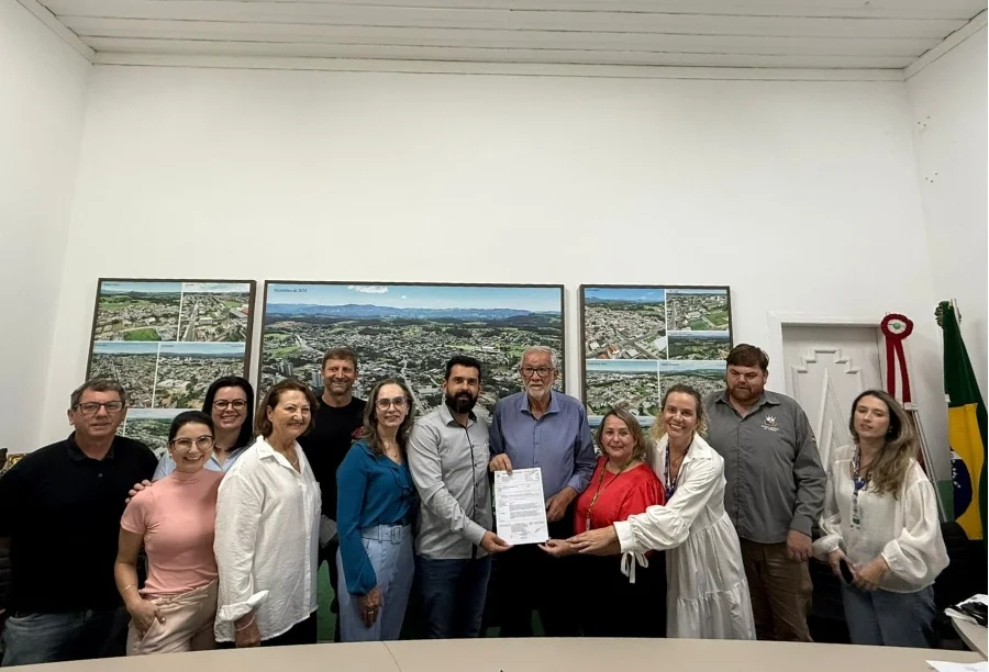 Prefeito Ademir Magagnin e autoridades de Cocal do Sul seguram documento de ordem de serviço para pacote de obras de R$ 4,3 milhões em habitação e infraestrutura. Ao fundo, mapas aéreos da cidade em gabinete municipal.