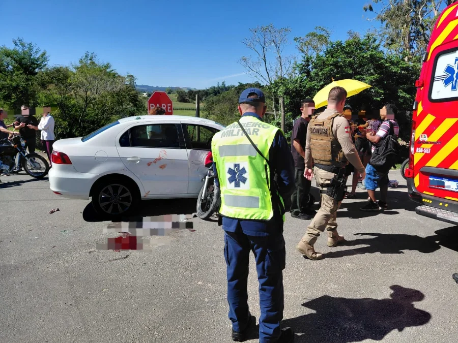 Acidente de trânsito em Morro da Fumaça envolvendo colisão lateral entre carro branco e motocicleta no bairro Estação Cocal. A imagem mostra bombeiro militar e policial militar atendendo a ocorrência próximo a uma ambulância.