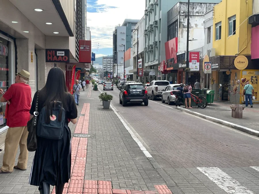 Rua movimentada no centro comercial de Criciúma com fachadas de lojas, pedestres caminhando e carros estacionados, ilustrando o movimento do varejo para as compras de Dia das Mães.