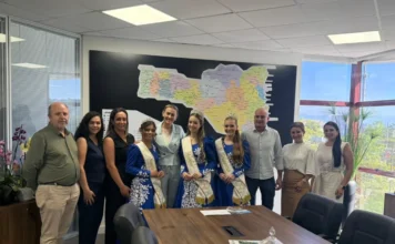 Comitiva de Meleiro promove VIII Agrimeleiro na capital Prefeito de Meleiro, Anderson Scardueli, e as soberanas da VIII Agrimeleiro em reunião com a secretária de Governo de Santa Catarina, Dani Pinheiro, em Florianópolis. A comitiva, vestindo trajes típicos azuis, posa para foto em frente a um mapa do estado durante a entrega do convite oficial da feira de agronegócio.