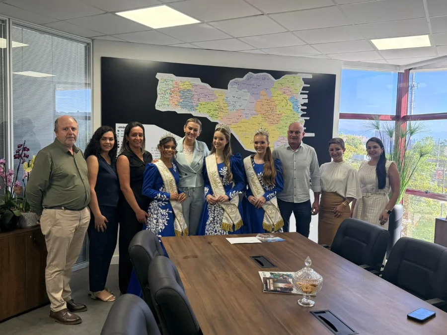 Prefeito de Meleiro, Anderson Scardueli, e as soberanas da VIII Agrimeleiro em reunião com a secretária de Governo de Santa Catarina, Dani Pinheiro, em Florianópolis. A comitiva, vestindo trajes típicos azuis, posa para foto em frente a um mapa do estado durante a entrega do convite oficial da feira de agronegócio.
