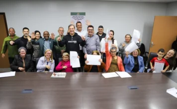 Criciúma acelera escrituras da Cohab antes de liquidação Grupo de moradores de Criciúma comemorando a entrega de escrituras da Cohab ao lado do prefeito Vagner Espindola em uma sala de reuniões na prefeitura. A imagem destaca a conquista da regularização fundiária urbana no município.
