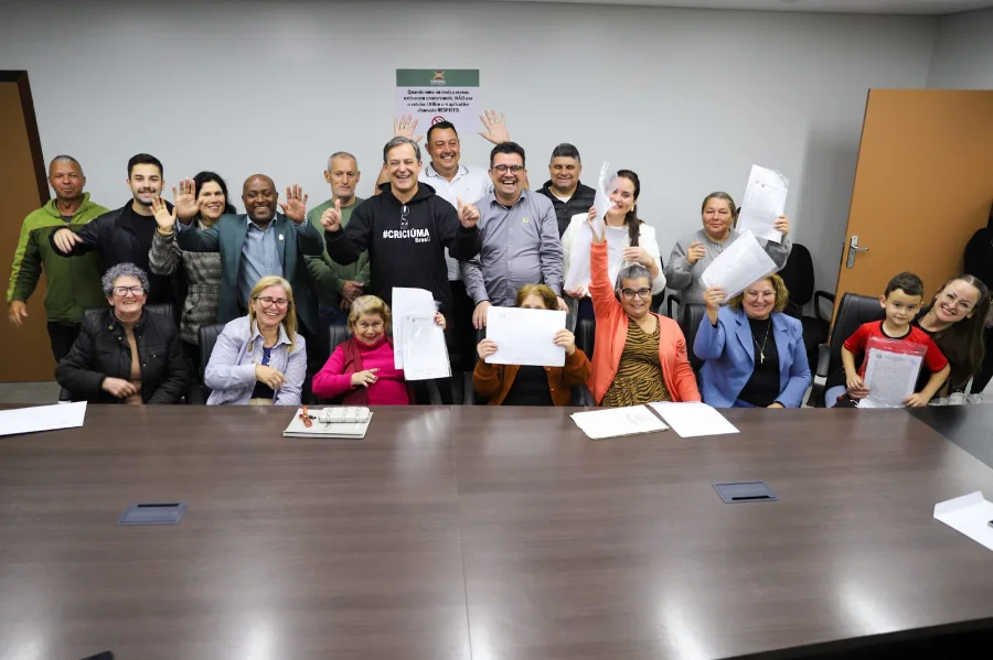 Grupo de moradores de Criciúma comemorando a entrega de escrituras da Cohab ao lado do prefeito Vagner Espindola em uma sala de reuniões na prefeitura. A imagem destaca a conquista da regularização fundiária urbana no município.