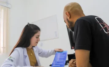 Criciúma alerta para alta de gripe e reforça vacinação Em uma Unidade Básica de Saúde (UBS) de Criciúma, uma profissional de saúde em jaleco branco explica informações sobre a prevenção da gripe e a importância da vacinação contra influenza a um cidadão de costas, entregando-lhe um folheto informativo azul sobre a mesa de consulta.