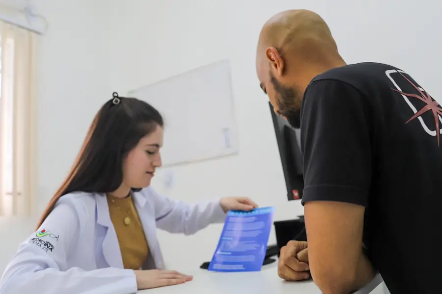 Em uma Unidade Básica de Saúde (UBS) de Criciúma, uma profissional de saúde em jaleco branco explica informações sobre a prevenção da gripe e a importância da vacinação contra influenza a um cidadão de costas, entregando-lhe um folheto informativo azul sobre a mesa de consulta.