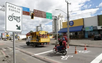 Criciúma amplia áreas de espera para motos em avenidas Motociclista utiliza nova área de espera sinalizada pela Prefeitura de Criciúma em cruzamento com semáforo. A cena mostra a sinalização horizontal com o ícone de motocicleta no asfalto e uma placa vertical indicativa, com um caminhão de manutenção da DTT ao fundo na Avenida Centenário.