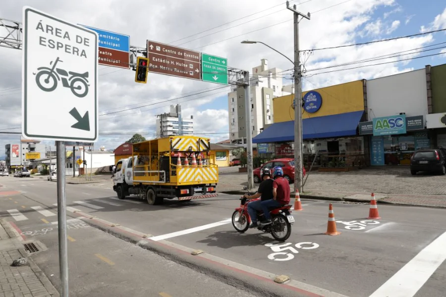 Motociclista utiliza nova área de espera sinalizada pela Prefeitura de Criciúma em cruzamento com semáforo. A cena mostra a sinalização horizontal com o ícone de motocicleta no asfalto e uma placa vertical indicativa, com um caminhão de manutenção da DTT ao fundo na Avenida Centenário.