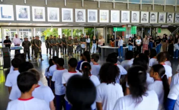 Criciúma celebra Dia do Hino Nacional com o Exército Apresentação da banda de música do 28º GAC do Exército Brasileiro no hall da Prefeitura de Criciúma. Militares em formação e uniformizados tocam instrumentos de sopro e percussão para uma multidão de estudantes da rede municipal e servidores no Paço Municipal durante ato cívico do Dia do Hino Nacional e Semana do Exército. Retratos históricos adornam a parede ao fundo.