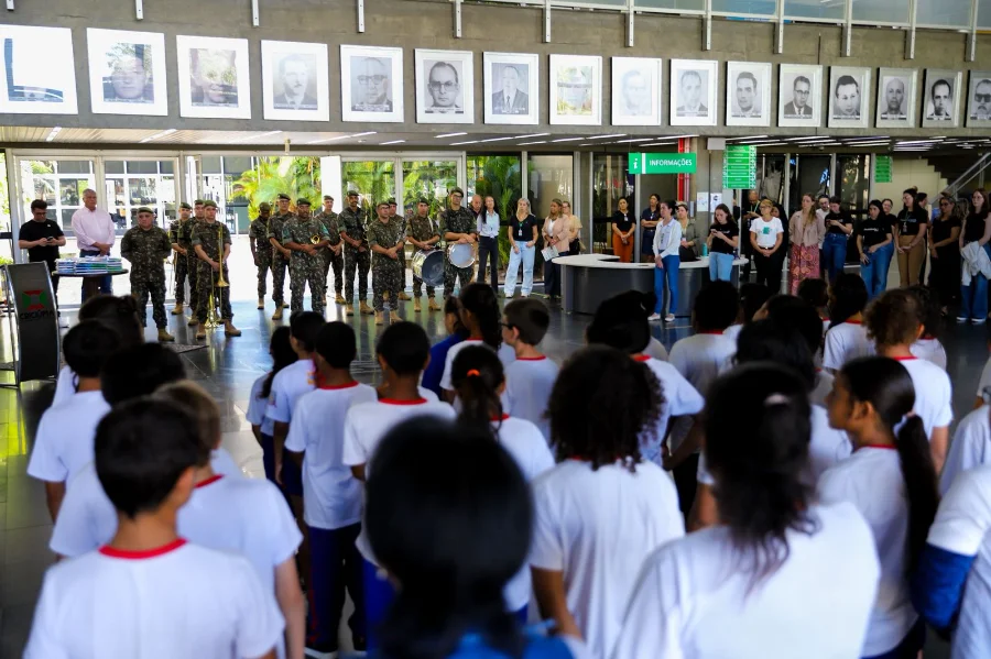 Apresentação da banda de música do 28º GAC do Exército Brasileiro no hall da Prefeitura de Criciúma. Militares em formação e uniformizados tocam instrumentos de sopro e percussão para uma multidão de estudantes da rede municipal e servidores no Paço Municipal durante ato cívico do Dia do Hino Nacional e Semana do Exército. Retratos históricos adornam a parede ao fundo.