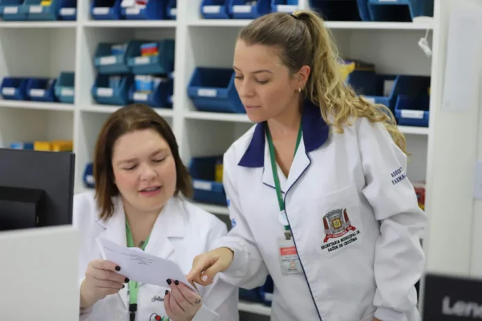 Criciúma é selecionada para projeto nacional do SUS Duas profissionais da Secretaria Municipal de Saúde de Criciúma, vestindo jalecos brancos, analisam um documento técnico em uma farmácia municipal. Ao fundo, prateleiras organizadas com insumos médicos representam a estrutura de atendimento do SUS em Criciúma, destacando a qualificação e a educação permanente em saúde.