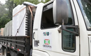 Criciúma entrega mobília nova para centros comunitários Foto de um caminhão da Prefeitura de Criciúma carregando mobiliário e equipamentos para Centros Comunitários. Na carroceria, estão visíveis pilhas de cadeiras plásticas brancas e grandes eletrodomésticos, como refrigeradores ou freezers, prontos para a entrega. O caminhão possui o logotipo oficial de Criciúma.