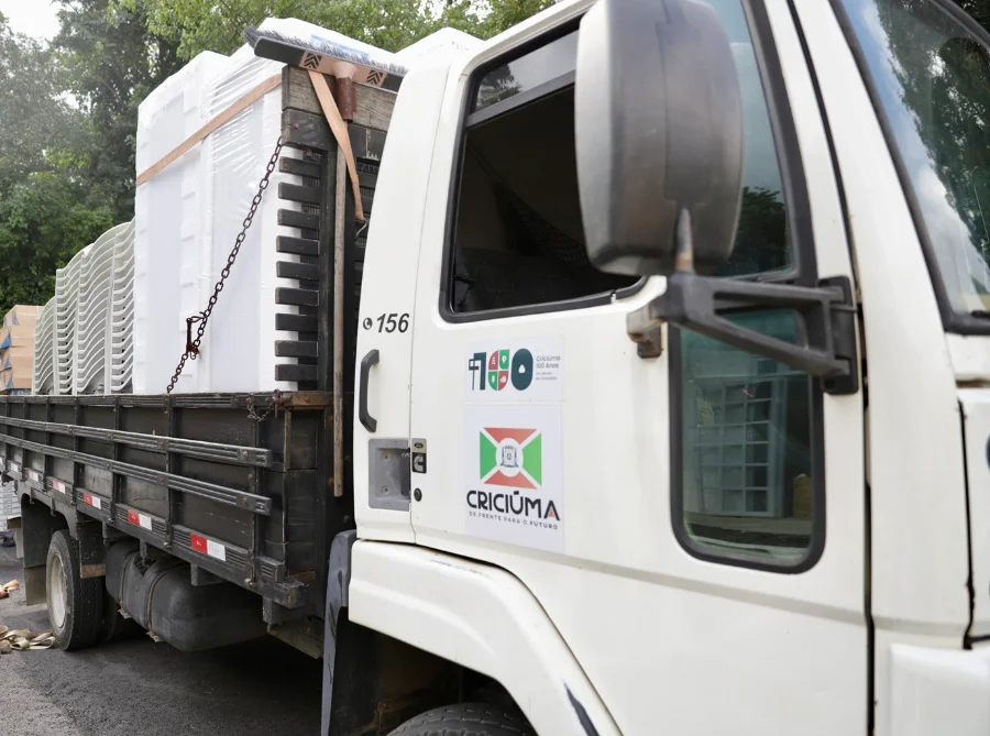 Foto de um caminhão da Prefeitura de Criciúma carregando mobiliário e equipamentos para Centros Comunitários. Na carroceria, estão visíveis pilhas de cadeiras plásticas brancas e grandes eletrodomésticos, como refrigeradores ou freezers, prontos para a entrega. O caminhão possui o logotipo oficial de Criciúma.