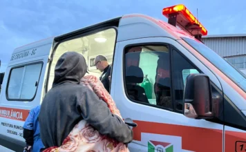 Criciúma interna seis pessoas com dependência no Pinheirinho Ambulância da Prefeitura de Criciúma em operação de internação de pessoas com dependência química no bairro Pinheirinho. A imagem mostra um profissional de saúde auxiliando uma pessoa com cobertor a entrar no veículo para transporte médico.