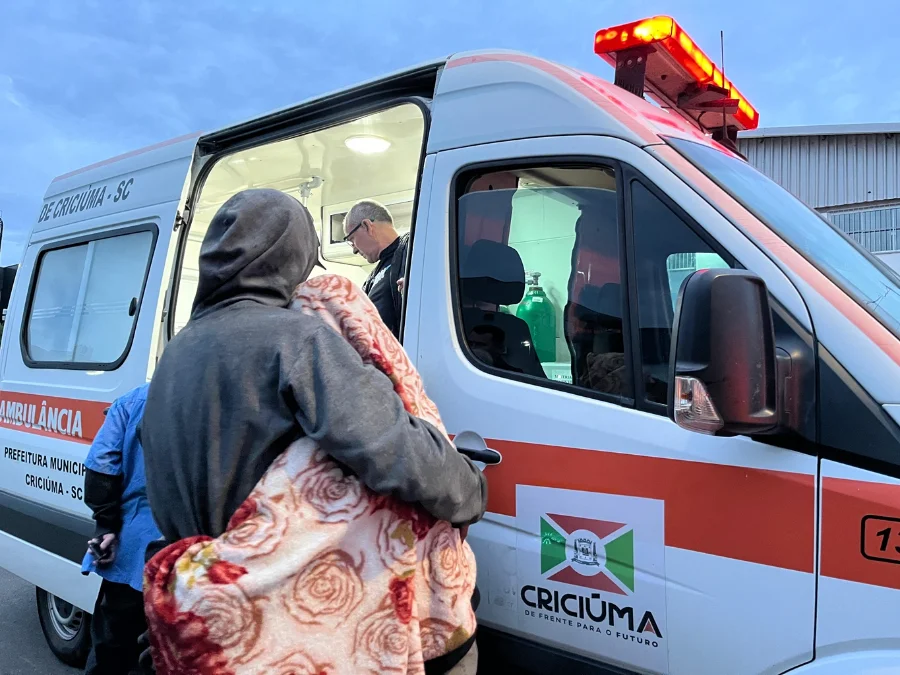 Ambulância da Prefeitura de Criciúma em operação de internação de pessoas com dependência química no bairro Pinheirinho. A imagem mostra um profissional de saúde auxiliando uma pessoa com cobertor a entrar no veículo para transporte médico.