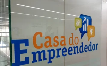 Criciúma lança programa Avança MEI para acelerar negócios Entrada de vidro da Casa do Empreendedor em Criciúma, local de apoio e capacitação para microempreendedores, incluindo iniciativas como o programa Avança MEI. O logotipo colorido apresenta ícones de engrenagem, gráfico de crescimento e lâmpada sobre texto azul e laranja. O interior claro e moderno do escritório é visível através do vidro.