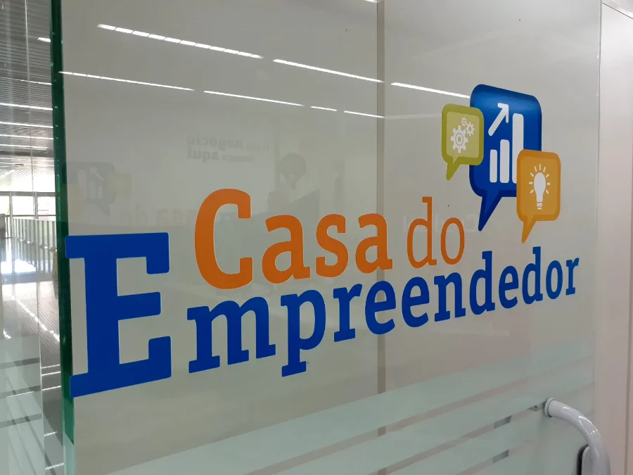 Entrada de vidro da Casa do Empreendedor em Criciúma, local de apoio e capacitação para microempreendedores, incluindo iniciativas como o programa Avança MEI. O logotipo colorido apresenta ícones de engrenagem, gráfico de crescimento e lâmpada sobre texto azul e laranja. O interior claro e moderno do escritório é visível através do vidro.