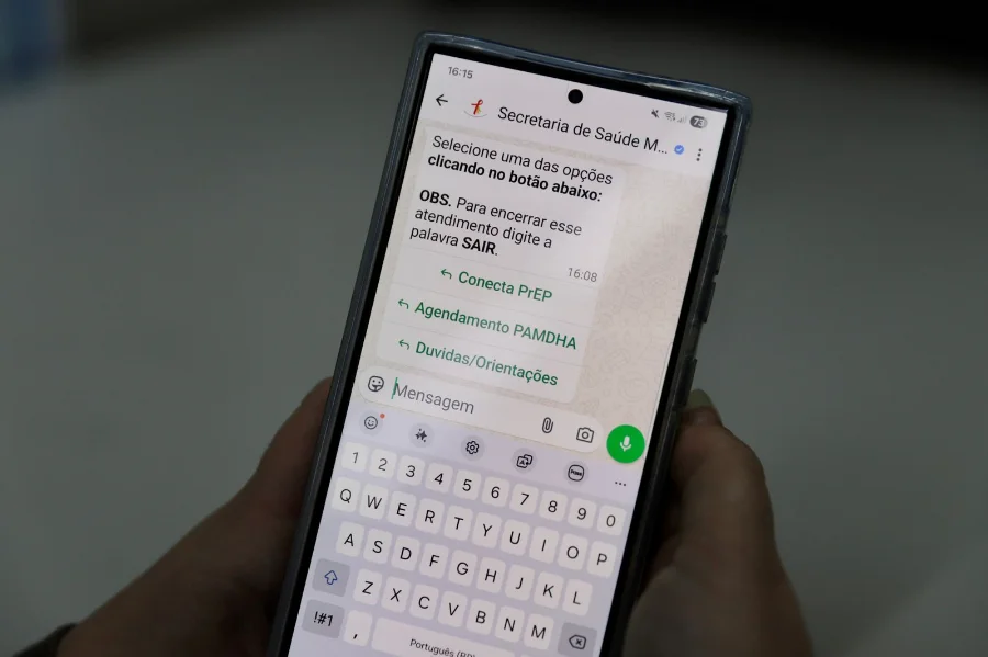 Pessoa segurando smartphone com a tela do WhatsApp aberta no serviço Conecta PrEP da Secretaria de Saúde de Criciúma. Na tela, aparecem opções de menu para agendamento e orientações sobre prevenção ao HIV via teleatendimento.