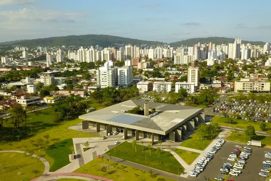 Vista aérea do Paço Municipal de Criciúma e do centro urbano, destacando a sede administrativa da prefeitura e prédios ao fundo sob céu claro. A imagem ilustra a infraestrutura da cidade líder em gestão municipal em Santa Catarina.