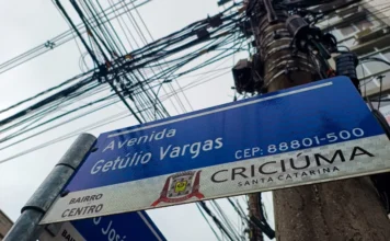 Limpa Fios: trânsito terá alterações em Criciúma nesta terça Vista inferior de uma placa de rua azul na Avenida Getúlio Vargas, no Centro de Criciúma, ao lado de um poste de utilidade pública com um emaranhado denso e desordenado de fiação aérea e cabos obsoletos, exemplificando o problema combatido pela Operação Limpa Fios.