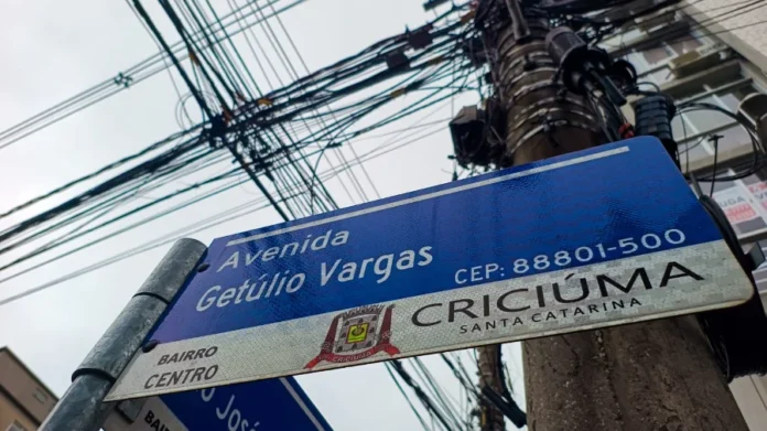 Criciúma realiza nova etapa da operação Limpa Fios Vista inferior de uma placa de rua azul na Avenida Getúlio Vargas, no Centro de Criciúma, ao lado de um poste de utilidade pública com um emaranhado denso e desordenado de fiação aérea e cabos obsoletos, exemplificando o problema combatido pela Operação Limpa Fios.