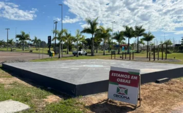 Criciúma terá novos chafarizes em dois parques urbanos Vista da base circular de concreto em construção do novo chafariz interativo no Parque dos Imigrantes em Criciúma, com placa de sinalização de obras da Prefeitura Municipal em destaque e palmeiras ao fundo.