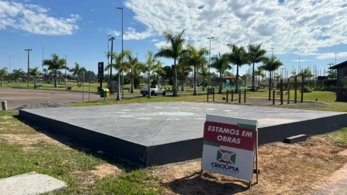 Criciúma terá novos chafarizes em dois parques urbanos Vista da base circular de concreto em construção do novo chafariz interativo no Parque dos Imigrantes em Criciúma, com placa de sinalização de obras da Prefeitura Municipal em destaque e palmeiras ao fundo.