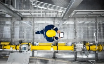 Custo de matéria-prima dispara com guerra no Oriente Médio Trabalhador industrial com capacete amarelo utiliza tablet para monitorar operações em fábrica com tubulações, representando a gestão da produção e o impacto da alta nos custos de matéria-prima na indústria.