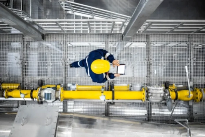 Custo de matéria-prima dispara com guerra no Oriente Médio Trabalhador industrial com capacete amarelo utiliza tablet para monitorar operações em fábrica com tubulações, representando a gestão da produção e o impacto da alta nos custos de matéria-prima na indústria.